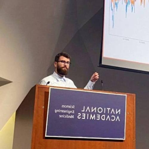 Joseph Wittrock stands behind a lectern while presenting. A screen displaying a line graph is over his shoulder.